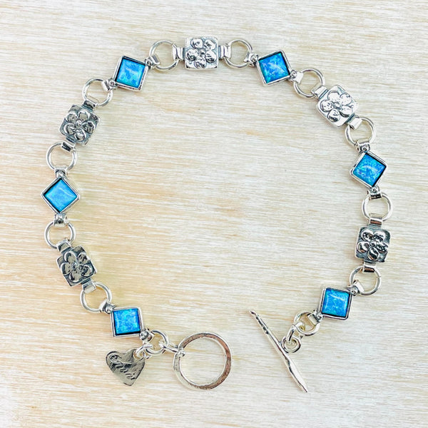 Six bright blue square opal links are spaced along the bracelet in a regular pattern of opal link, silver circle, silver squre link with flower design, circle, then opal again. Little silver heart charm at the end, with a T bar catch.