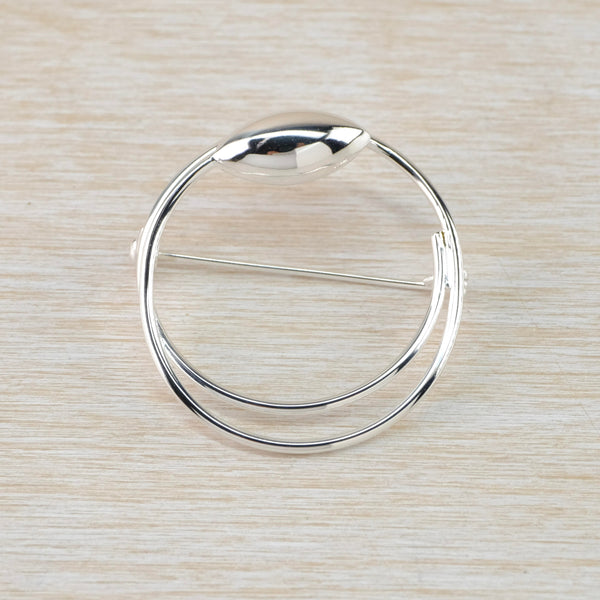 A high polished round outline silver brooch. At the top is a shiny silver pod shape and the bottom half has two silver strands