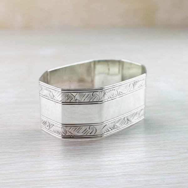 Octagonal silver napkin ring. Top and bottom is an engraved stripe with dashes and shading finished with 2 horizontal lines. The middle section is plain polished silver.