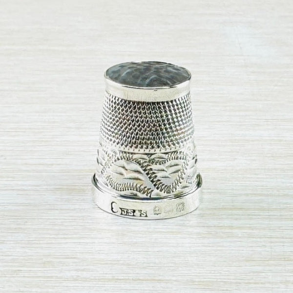 Silver thimble with a dark coloured top made from agate. The top half of the thimble has repeated little dots, the bottom half has a swirl design that looks a little like a zip with semi circle markings behind. The bottom band is plain shiny silver with a visisble hallmark.