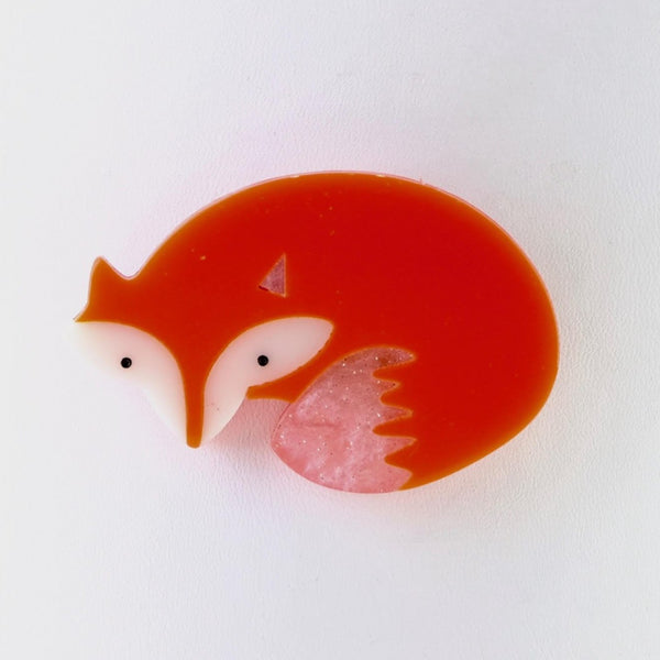 A simple fox shape, head at one end, curved body shape and a tail almost tucked in under his chin. It almost forms a circle. Fox orange in colour, the face has white cheeks, little black dot eyes and pointed ears. There is white shading at the end of his tail.