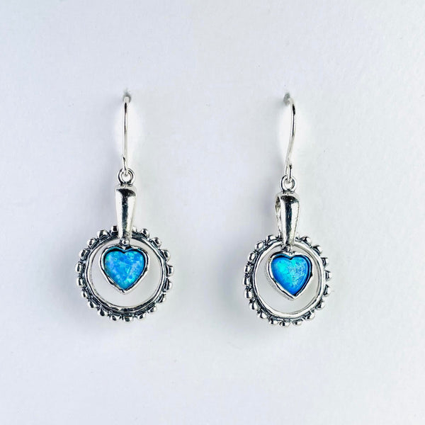 Drop earrings with a patterned silver circle  below a plain silver stalks. Hanging from the stalk within the circle is a bright blue, heart shaped opal surrounded in silver.