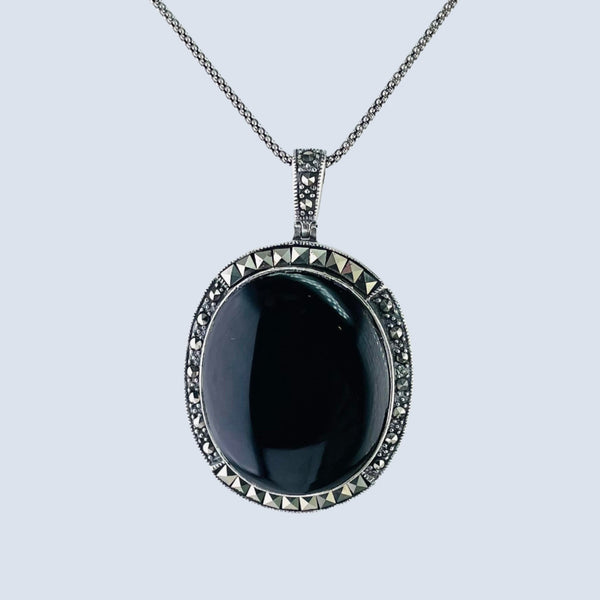 A shiny black oval onyx stone is surrounded by little marcasite stones cut in two shapes. The stones at the top and bottom are square cut, the ones on the side are circular. The pendant is attached to a marcasite set bail and is hanging off an oxidized chain.