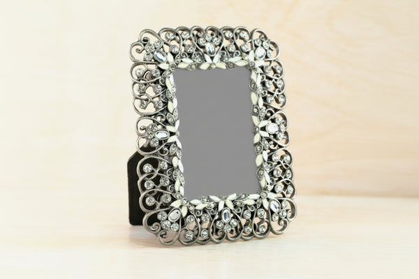 An ornate rectangular picture frame with a silver filigree border and clear rhinestones, standing on a light wooden surface against a pale background.
