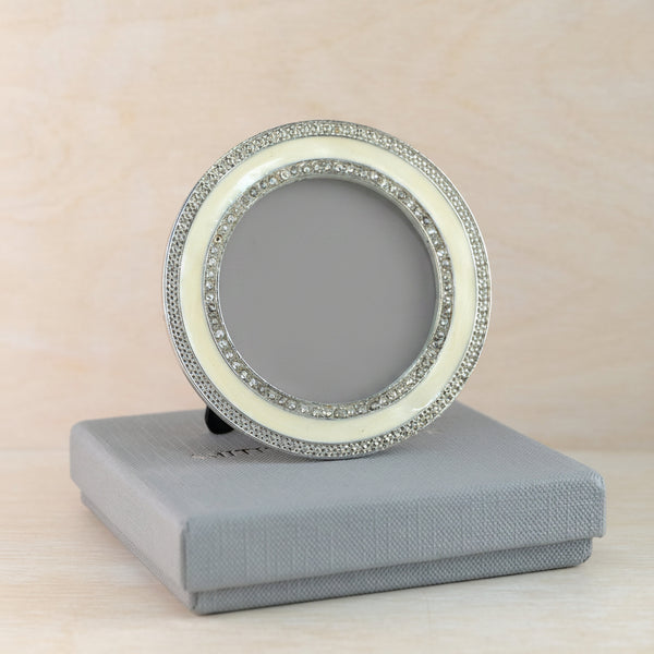 A round silver-toned picture frame with decorative crystal-like borders, leaning on a gray presentation box against a light wood background.