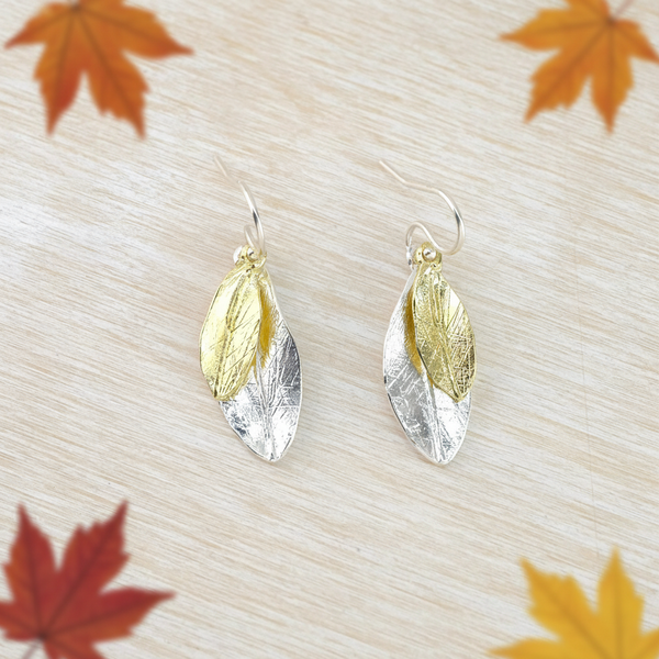 A pair of gold-plated and silver earrings designed to resemble dangling leaves, displayed on a white wooden surface.