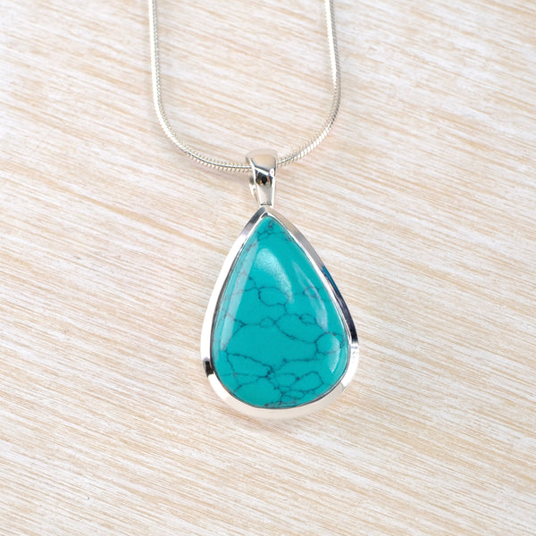 Turquoise teardrop pendant on a silver chain against a light wood background