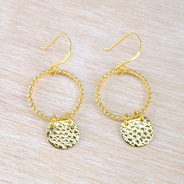Gold hoop earrings with round disc pendants on a light wood surface