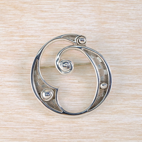 Silver circular brooch with a swirl design on a beige woven background