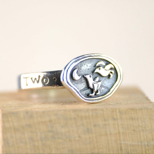 Silver ring with design of two birds and a gold crescent moon on a wooden block against a light background