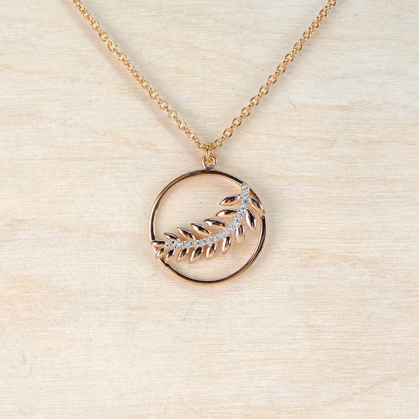 Gold necklace with a circular pendant  with a fern motif within on a beige background