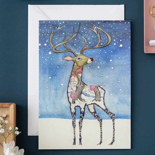 A Christmas greeting card featuring a watercolor illustration of a reindeer with a glowing red nose, antlers adorned with snow, and a winter landscape in the background. The card is displayed with a white envelope, against a navy background.