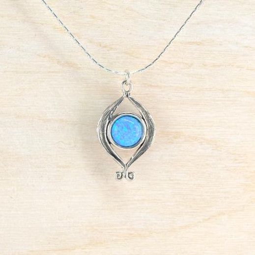 A silver pendant necklace featuring a round, vibrant blue opal-like gemstone surrounded by a textured, organic silver frame, suspended from a delicate silver chain on a light wood background.
