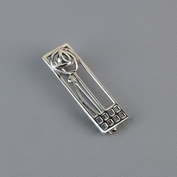 A silver rectangular brooch, with a stylised rose and geometric elements, on a grey background