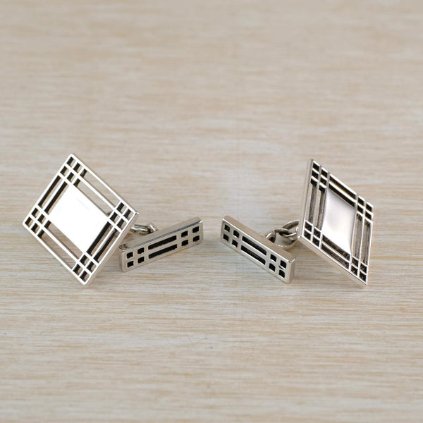 A pair of silver rectangular cufflinks with geometric frame designs and  displayed on a light wood surface.