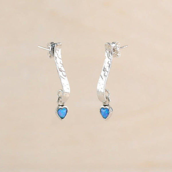 A pair of silver huggie hoop earrings with textured curved bands and dangling heart charms, displayed  against a soft beige background.