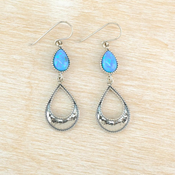 A pair of silver dangle earrings with teardrop-shaped light blue opal gemstones and  a textured silver teardrop  displayed on a light wood grain background.