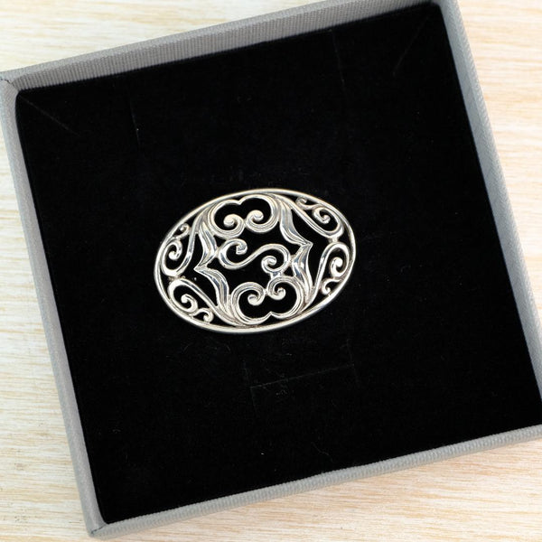 An oval silver filigree brooch with intricate scrollwork, displayed inside a black velvet-lined gray gift box on a light wood surface.
