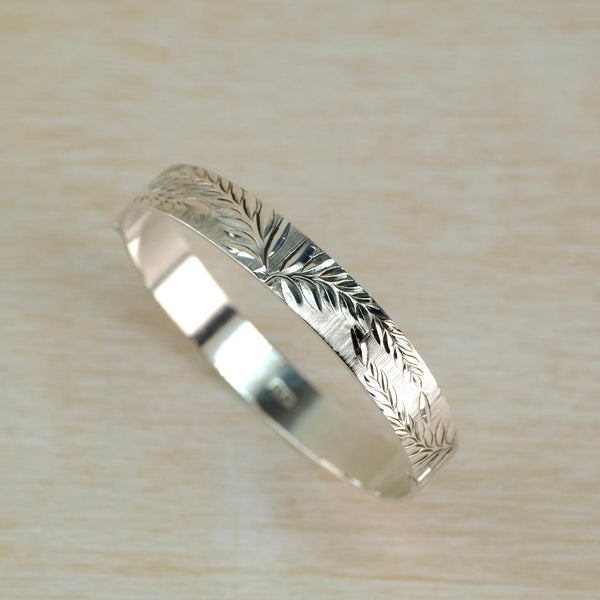 A polished silver bangle bracelet with engraved leaf patterns, resting on a light beige wooden surface.