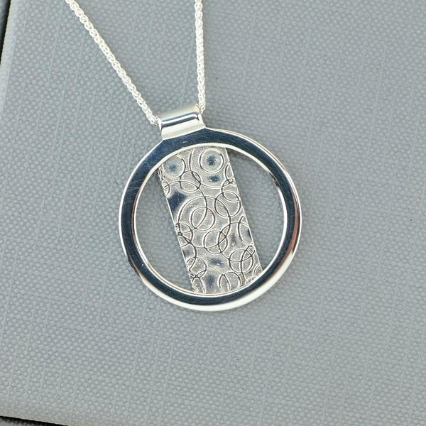 A silver pendant necklace with a circular frame and a textured rectangular center, displayed on a gray textured jewelry box.