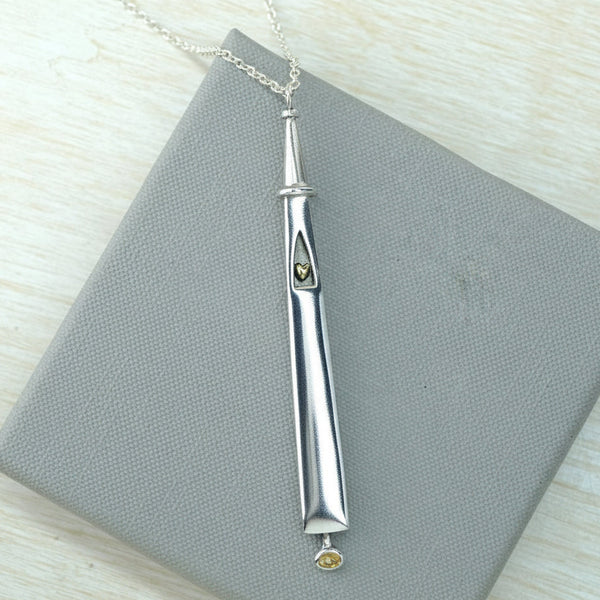 A silver pendant necklace  with a little gold heart and a citrine dangling at the bottom, displayed on a grey box.