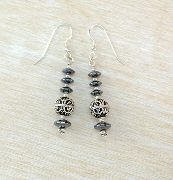 A pair of silver dangle earrings with dark gray beads and a central filigree sphere, displayed on a light wood background.