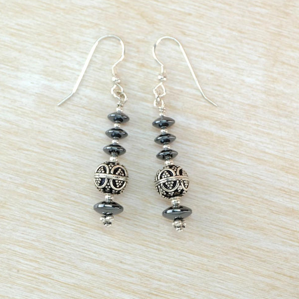 A pair of silver dangle earrings with dark gray beads and a central filigree sphere, displayed on a light wood background.