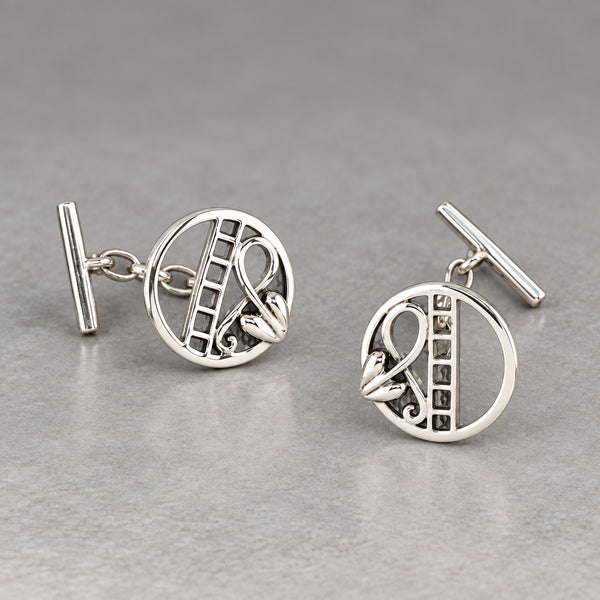 A pair of silver round cufflinks with a cutout floral and scroll design, displayed on a textured gray surface