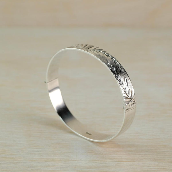 A polished silver hinged bangle bracelet with engraved leaf patterns, resting on a light wood surface.