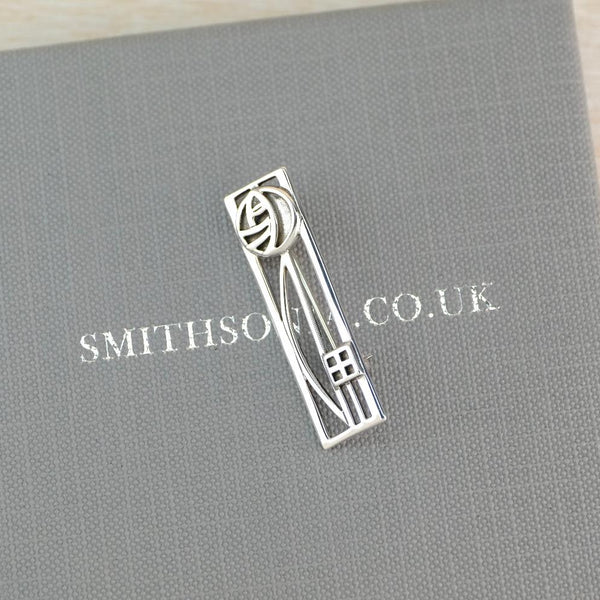 A silver Arts and Crafts style  brooch with abstract floral and geometric design, displayed on a textured gray Smithsonite gift box.