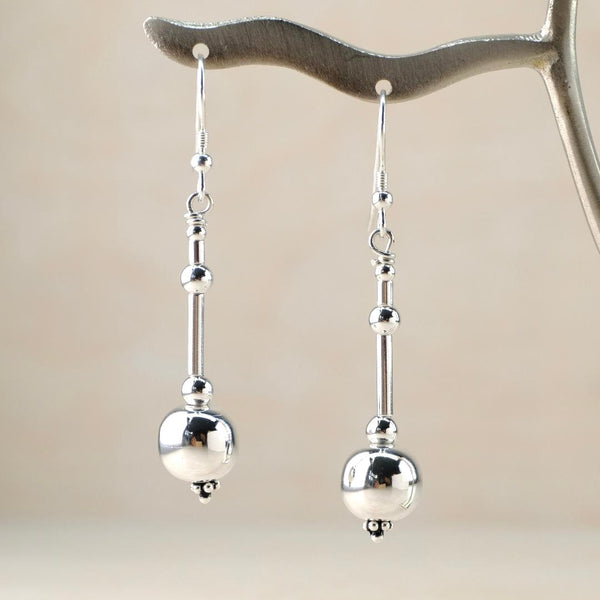A pair of dangly earrings on a silver hook. Each earring is composed of a series of beads.  At the bottom is a spherical smooth and shiny bead 1cm in diameter, above that there are two smaller spheres alternating with two slender cylindrical beads in a balanced design.