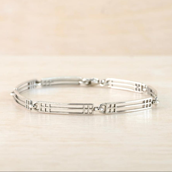 A polished silver  bracelet with a geometric, multi-barred  design, resting on a light beige wooden surface.