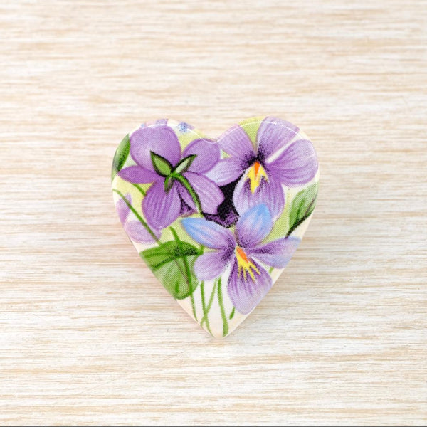 A flat  heart-shaped ceramic brooch which has been transfer printed with a pretty pattern of violet flowers and leaves in a vintage style and colourway. On a cream background there are 3 violet flowers with subtle green leaves 
