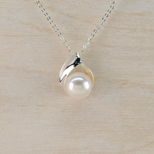 A silver necklace with a leaf-shaped pendant holding a single white pearl, displayed on a light wood background.