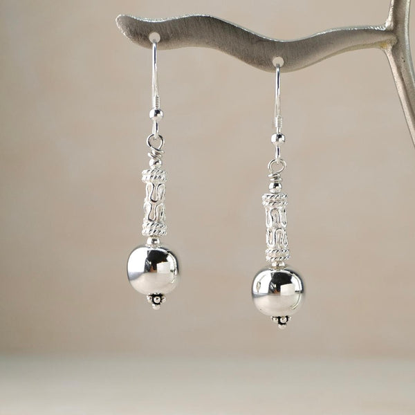 a pair of dangly earrings with a hook fitting. A super shiny and smooth spherical silver bead below a cylindrical bead which is delicately decorated with scrolls and dots and has some texture.