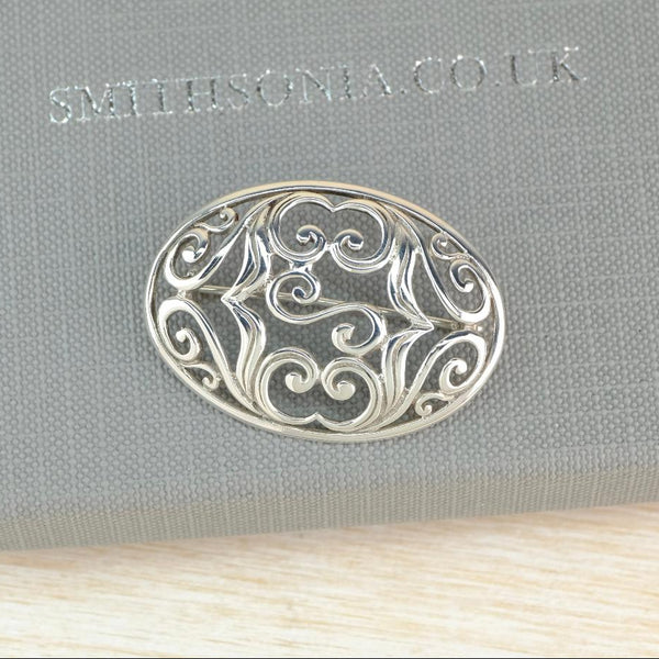 An oval silver filigree brooch with intricate scrollwork, displayed on a textured gray fabric box.