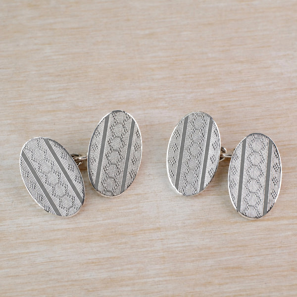 A pair of oval cufflinks made from silver, with intricate engine-turned geometric engraving displayed on a light wood background.