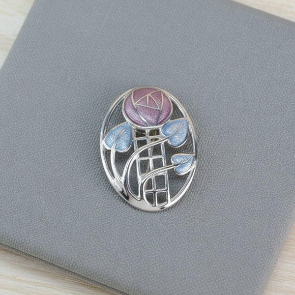 An oval silver brooch featuring a floral design with a pink rose, blue leaves, and a ladder motif, displayed on a textured gray box.