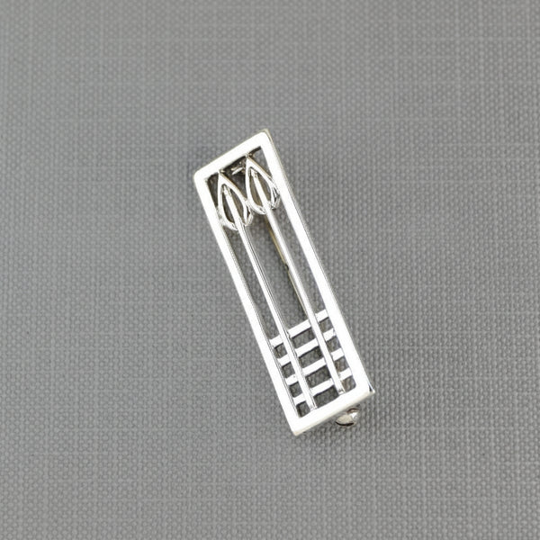 A silver lapel pin or brooch shaped like a stylized window with geometric and leaf-like elements, displayed on a textured gray surface.