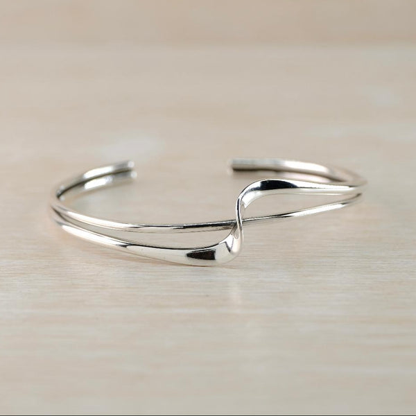 A polished silver open cuff bracelet with a twisted, flowing design, resting on a light beige wooden surface.