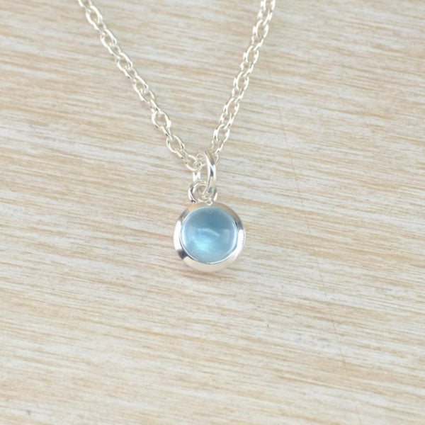 A small round silver and blue stoned pendant hanging from an oval linked silver chain. The pale blue smooth stone is set in a shiny round frame with a shiny silver ring for the chain to thread through.