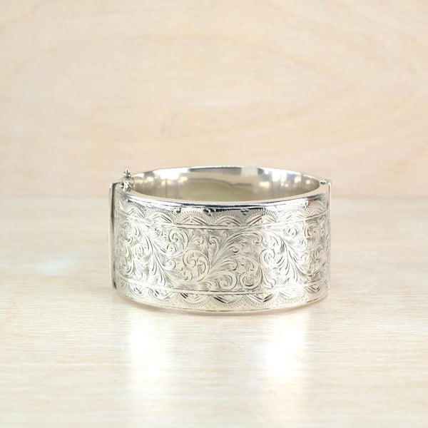 A wide silver cuff bracelet with an engraved floral scroll pattern, resting on a light wood surface.