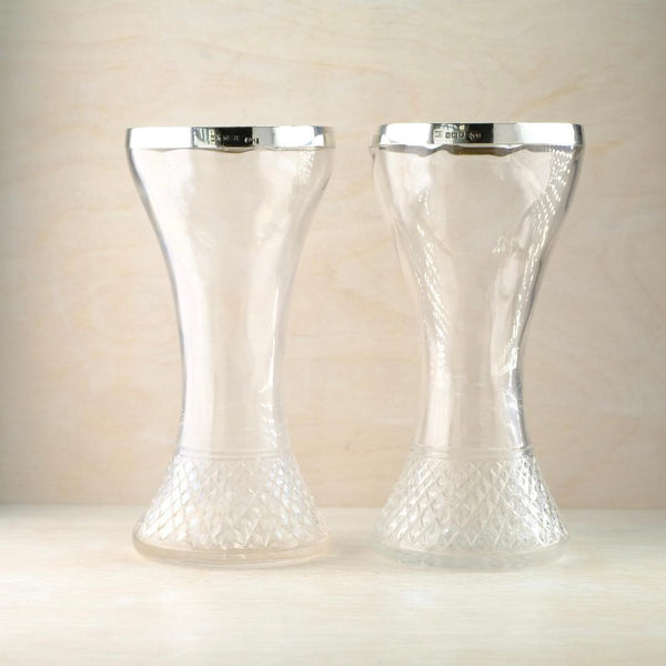 A pair of identical clear glass vases the rims of which are collared with shiny silver in a square section approximately 1 cm in depth. The silver collars both bear a clear silver hallmark. The glass vases  have a narrower 
 mid section like a waist and open up into a wider fluted shape. The repeated diamond shaped patterning is on the lower 5cm of  the vases .