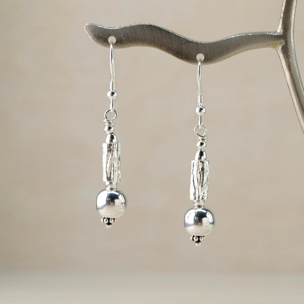 Dangly earrings made of different shiny silver beads hanging below the hook. On each earring there is a cylindrical bead with a  texture like tree bark and below that a spherical smooth ball bead.