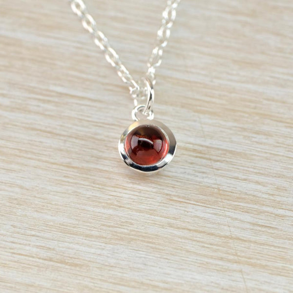 A small round silver and red stoned pendant hanging from a oval linked silver chain. The dark red smooth stone is set in a shiny round frame with a shiny silver ring for the chain to thread through. 