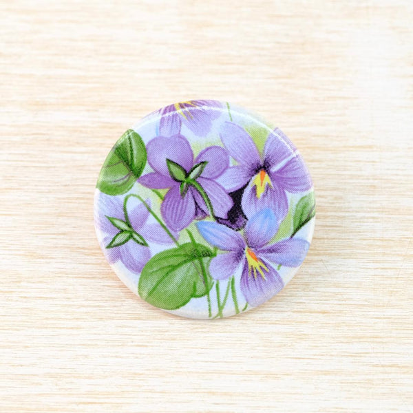 A flat round ceramic brooch which has been transfer printed with a pretty pattern of violet flowers and leaves in a vintage style and colourway. On a cream background there are 5 violet flowers with subtle green leaves 