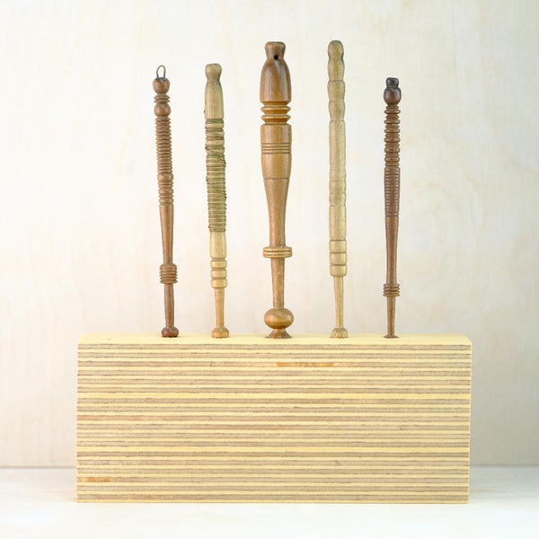 5 different wooden lace bobbins, all different shapes and patterns stand in a solid rectangular pale wood block with a horizonyal striped effect.