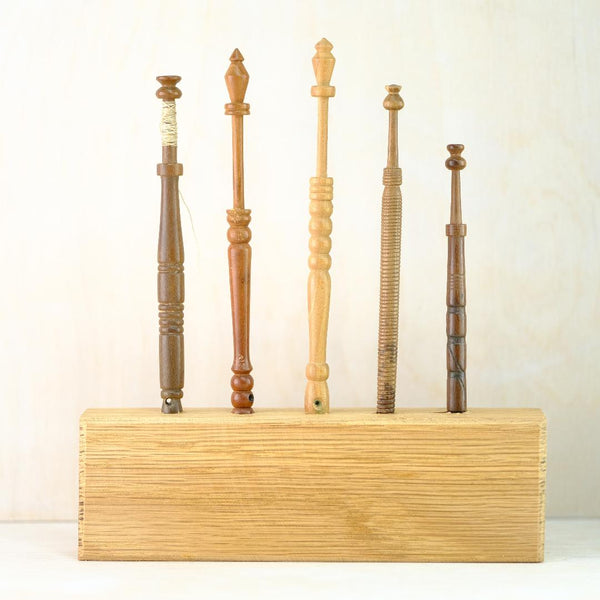 5 different wooden lace bobbins, all different shapes and patterns stand in a solid rectangular pale wood block.
