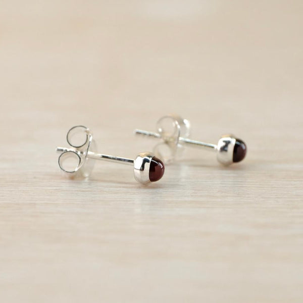 A pair of silver and red stoned stud earrings. A round smooth dark lred stone sits proud of a polished silver surround with a buttertly fitting on a silver post attached centrally.