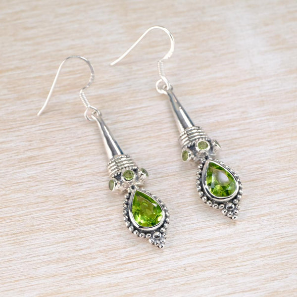 long silver and peridot earrings . Peridot is a fresh cheerful light green. A teardrop shaped and faceted pridot stone is set below a decorated half cone shape which a three smaller round peridots set around it 